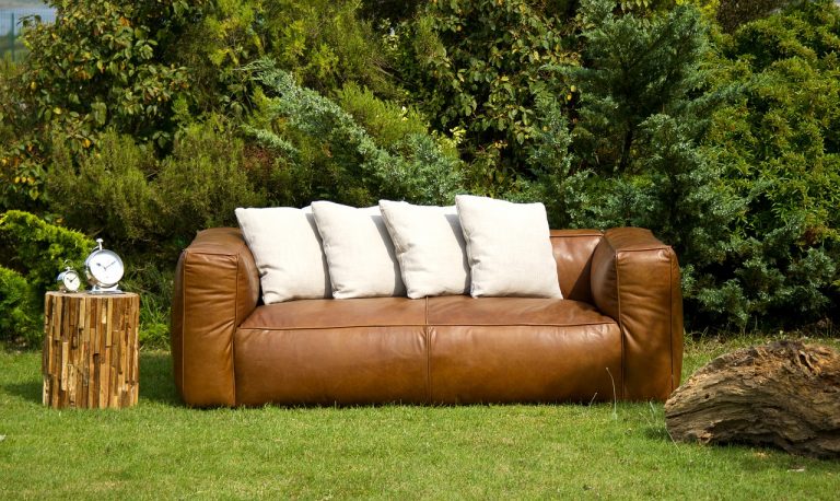 Auf einem Rasen steht ein braunes Kunstleder-Sofa, auf welchem vier helle Kissen drapiert sind. Daneben steht ein Beistelltisch, auf welchem sich zwei Wecker befinden.