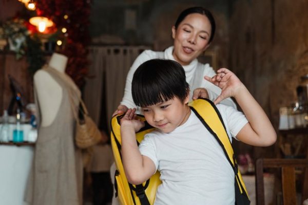 Einem Kind wird von einer Frau ein gelber Rucksack aufgesetzt