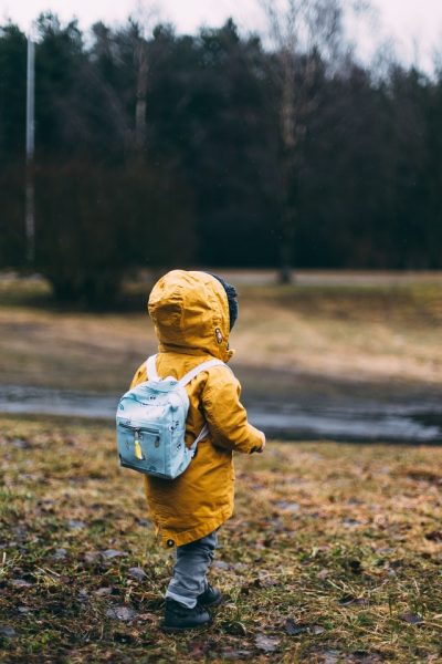 Ein Kind in einer gelben Regenjacke und mit einem kleinen Rucksack auf dem Rücken läuft auf einer Wiese vor einem Wald